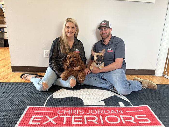 Owners Lexie and Chris sitting on the floor of their office with their two dogs and a mat in front of them with the Chris Jordan Exteriors logo on it